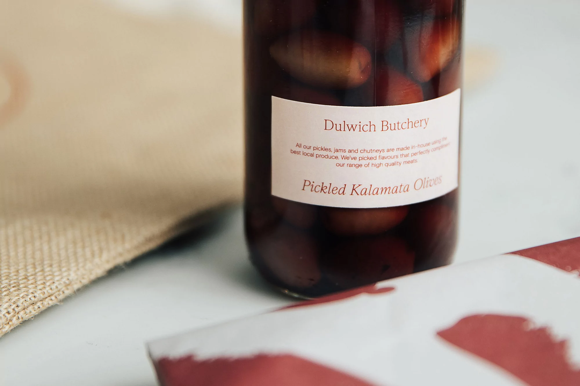 A jar of pickled Kalamata olives sits on a table with a hessian mat. The label reads 'Dulwich Butchery' and lists ingredients.