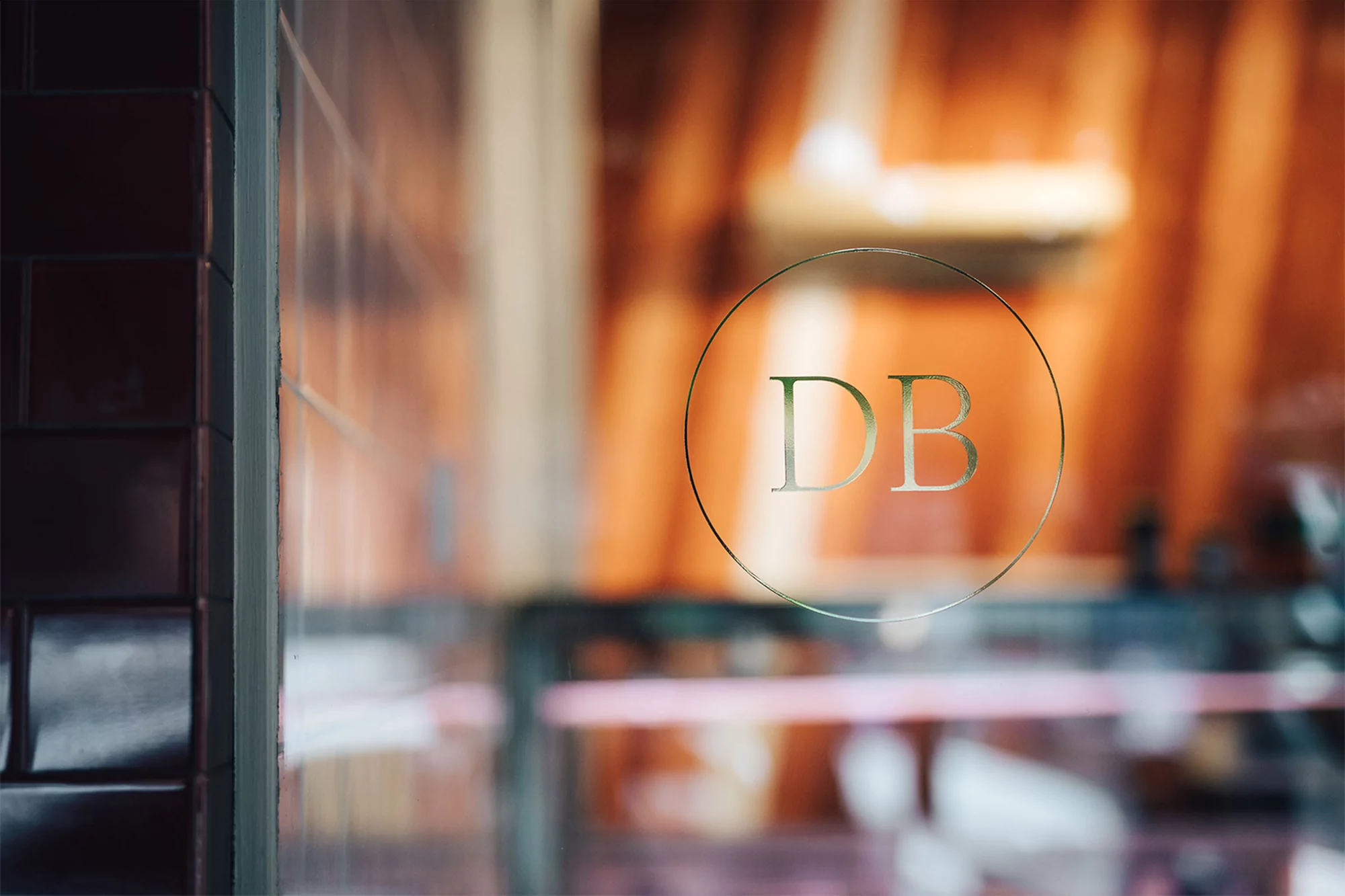 View through a glass door with the letters 'DB' in gold foil, revealing a blurred interior setting with warm, inviting lighting.
