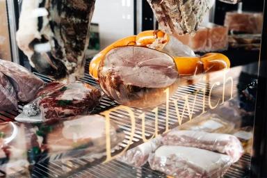 Close-up view of fresh raw cuts of meat and hams displayed inside a refrigerated retail display case with glass branding.