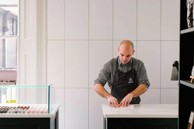 Adult male working at a white countertop in a food and beverage establishment, wearing an apron, with products in the background.