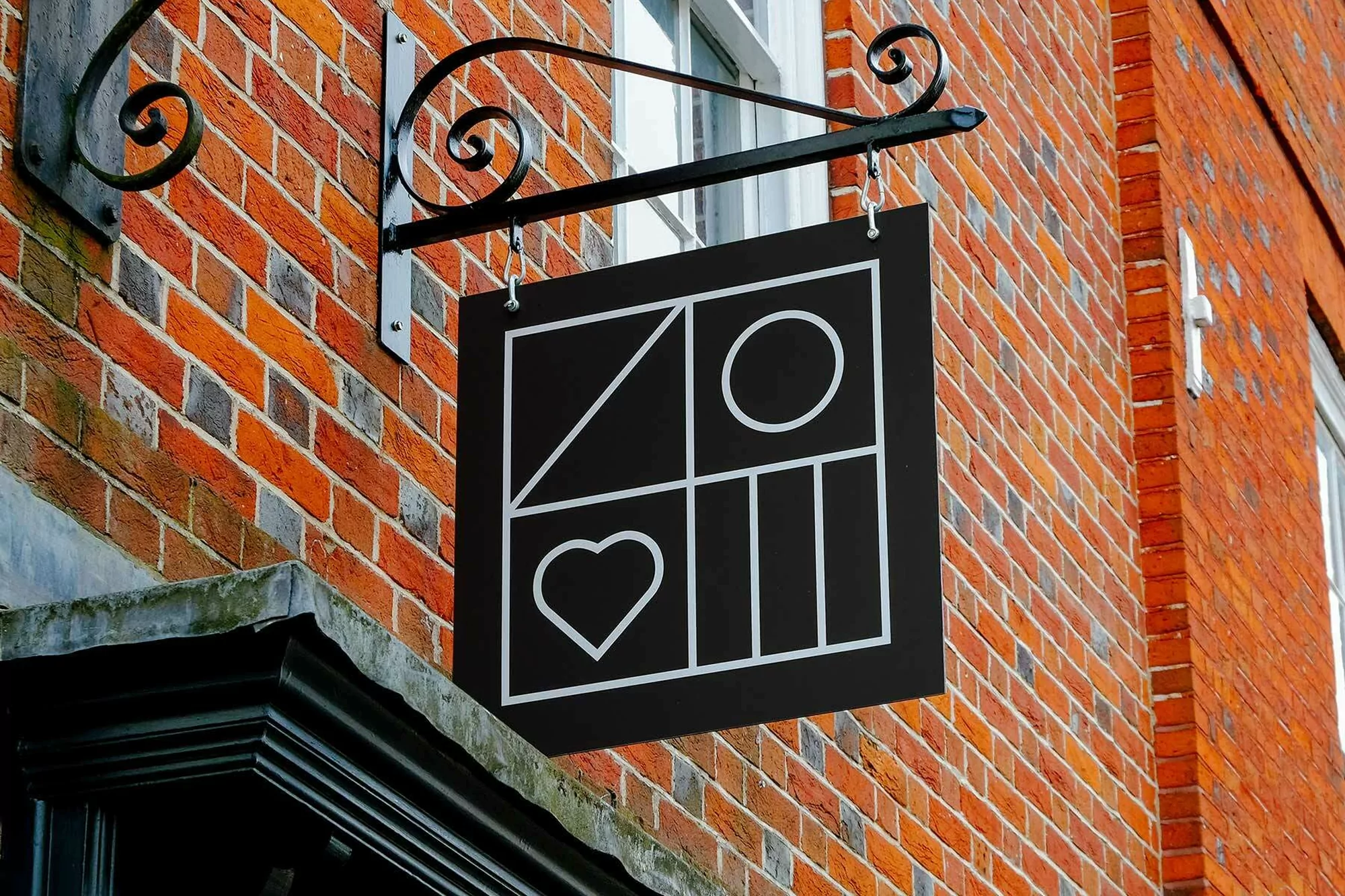 A black sign with white geometric shapes hangs on a wrought-iron bracket attached to an orange-red brick wall, with a window above.