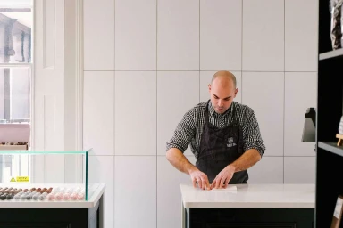 A bald man wearing a patterned shirt and dark apron preparing items at a white tiled counter for Hill St