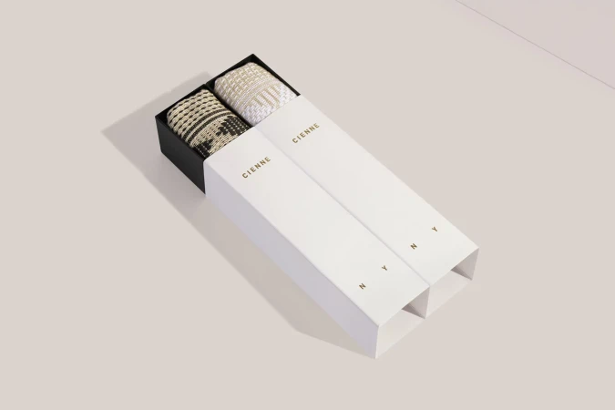Two boxes from Cienne NY containing woven textiles sit on a beige surface with bright, even lighting and sharp focus.