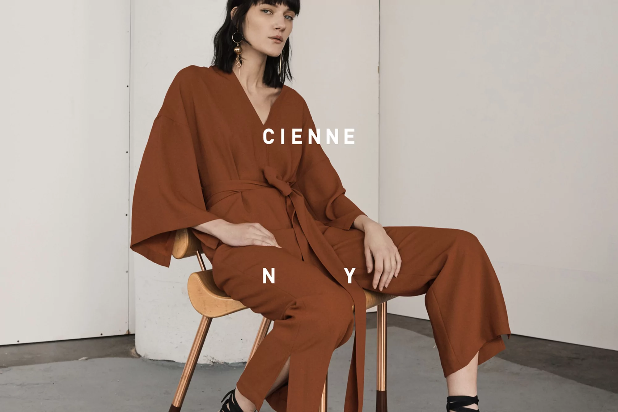 Adult female model in stylish brown jumpsuit sits on modern wood chair against white backdrop for fashion editorial photography.