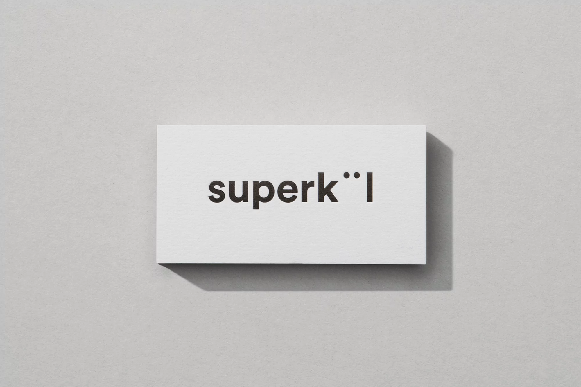 Promotional image of a white business card on a grey surface featuring the bold black text 'superk°°l', suggesting a modern brand.