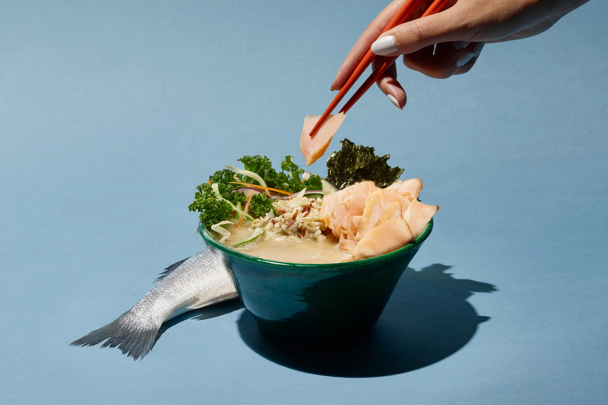 Ramen bowl presentation with fish, seaweed, greens, and broth, plus a hand holding chopsticks picking up a piece of meat, set on a blue surface.