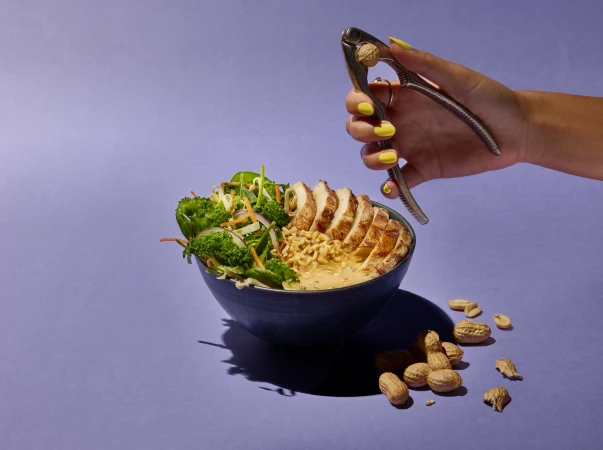 Chicken salad in a dark bowl with a nutcracker holding a peanut, against a lavender background. Food and beverage photo for Visie.