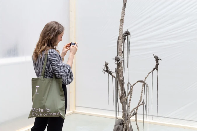 Woman in a patterned shirt takes a picture of a grey sculpture with her phone in a bright, minimalist art gallery setting.