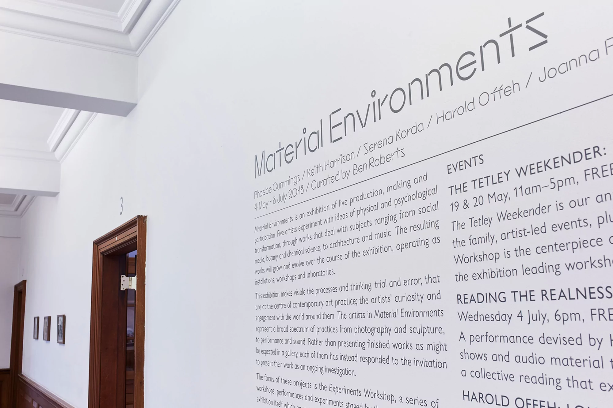Close-up shot of promotional material for the 'Material Environments' art exhibit featuring artist names, dates, and event details on white wall.
