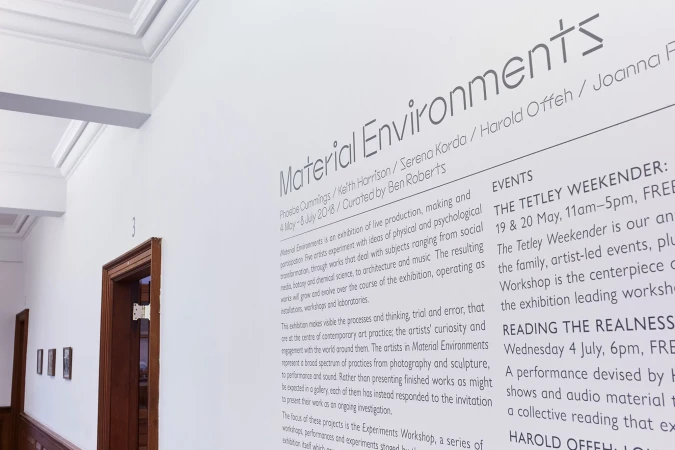 Close-up shot of promotional material for the 'Material Environments' art exhibit featuring artist names, dates, and event details on white wall.