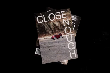 Stacked magazines with bold typography reading CLOSE ENOUGH featuring a toy car on the cover against a black background.