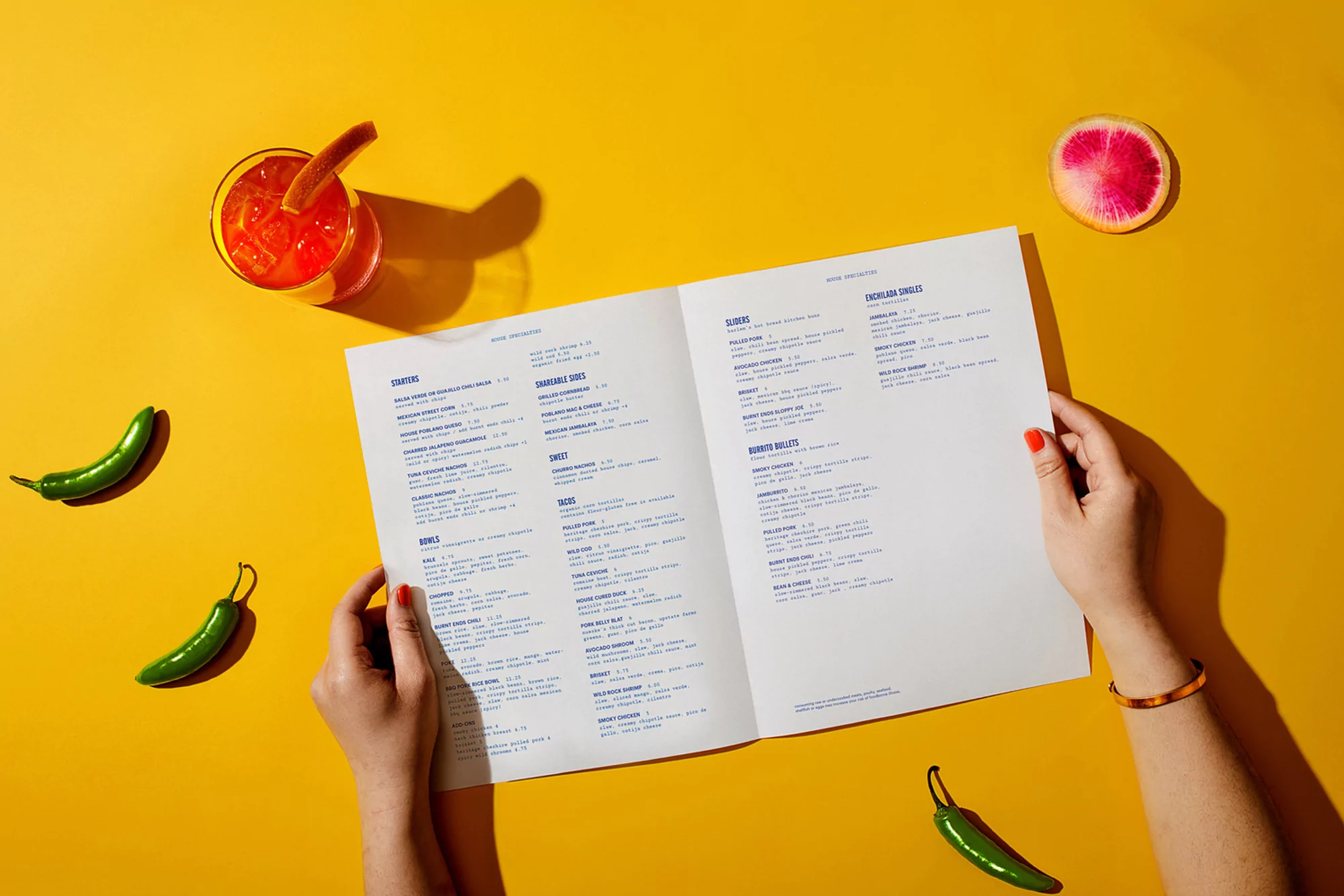 Overhead shot of a person holding a restaurant menu surrounded by a cocktail, jalapeños, and a radish against a yellow backdrop.