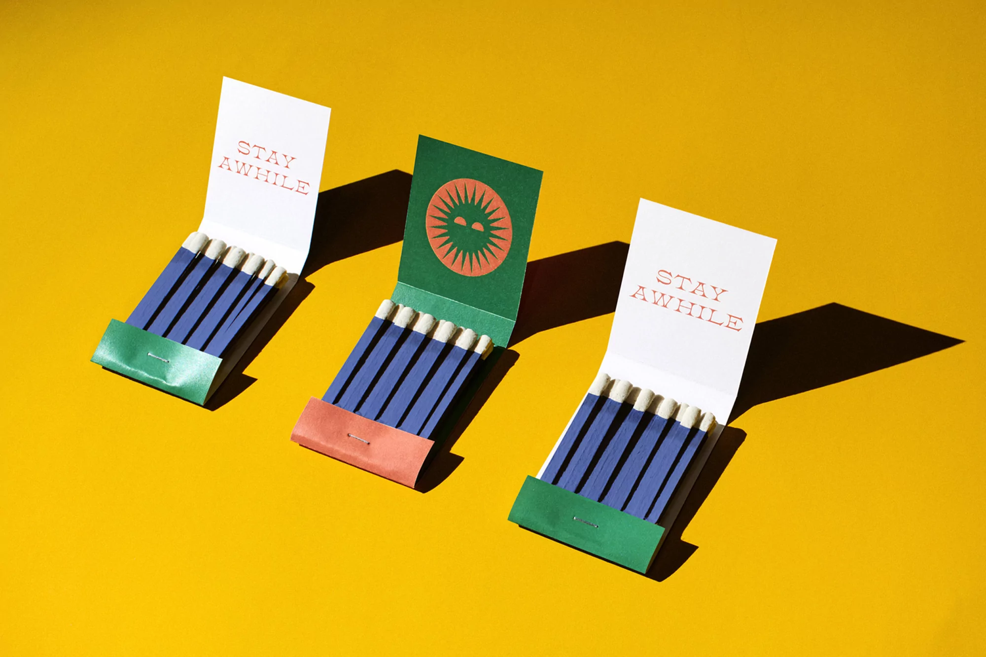 Three matchbooks displayed on a yellow surface, featuring the text 'Stay Awhile' and a stylized sun graphic, casting a soft shadow.