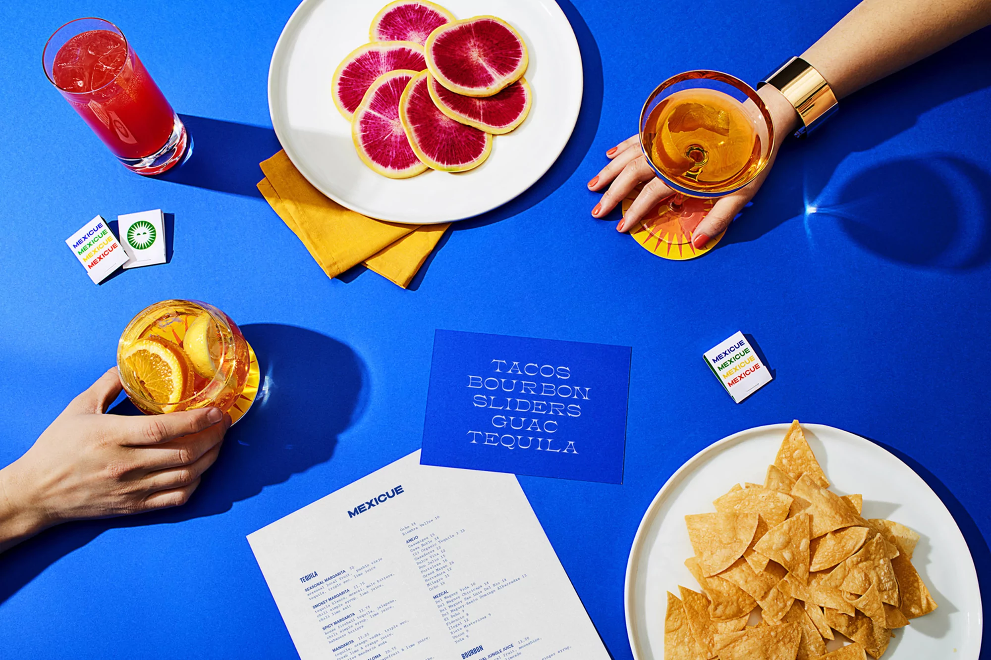 Overhead shot of bright blue table arrangement with plates of sliced citrus fruit, tortilla chips, and cocktails with menu and napkins.