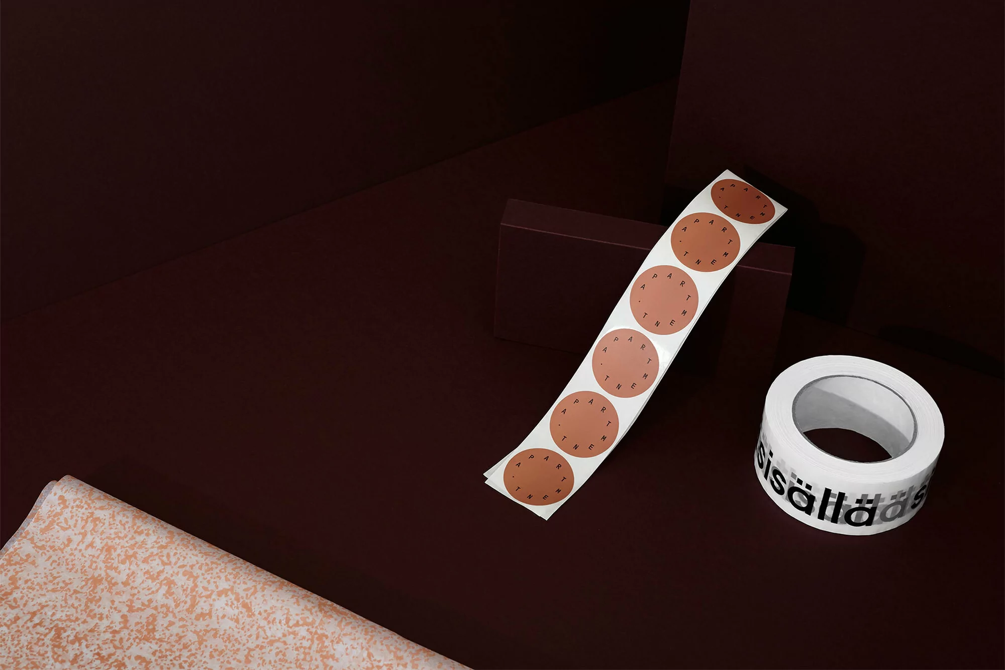 Still life of roll of round brown stickers, roll of tape with text, and folded patterned fabric on a dark reddish-brown surface.
