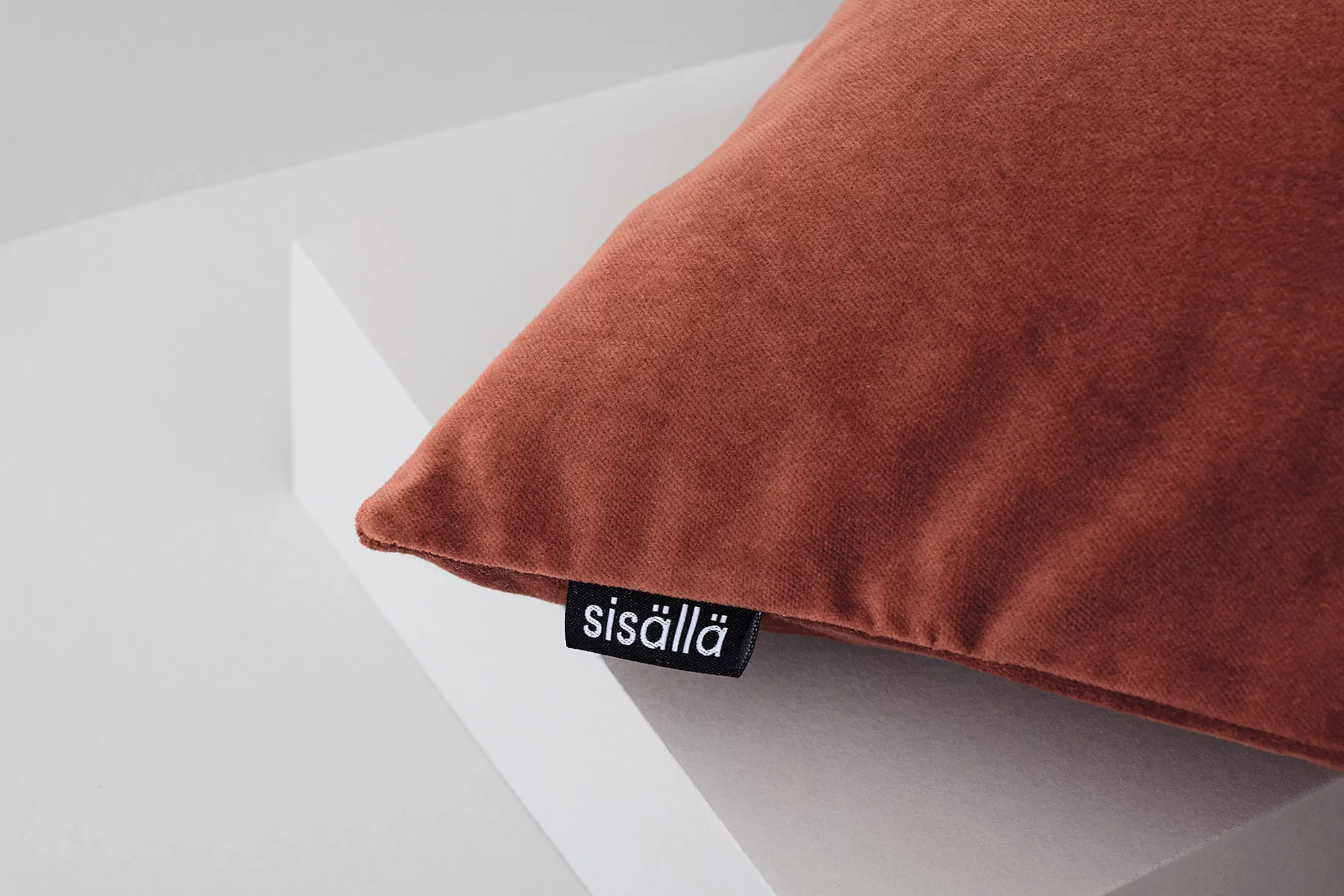 Close-up of a rusty-orange textured pillow against a white geometric backdrop with a black logo tag featuring white text.