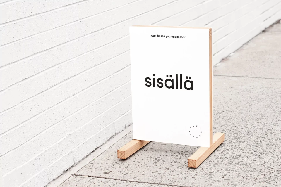 Minimalist sign with 'sisällä' in black letters, set against a white wall and light gray pavement; promotes a shop or event.