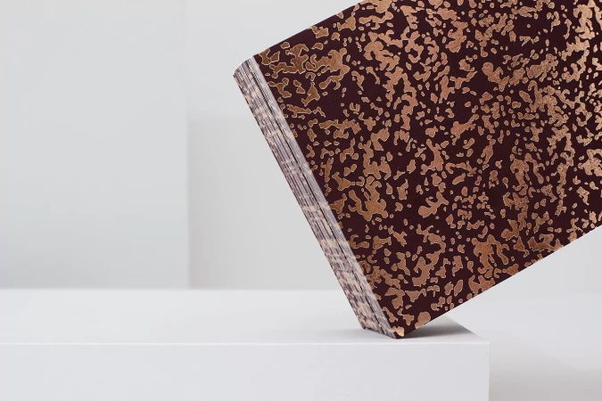 Stack of maroon paper with gold abstract pattern on a white surface, set against a plain white background, creating a minimalist composition.