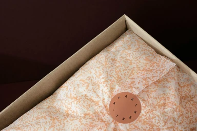 Cardboard box lined with peach patterned tissue paper sealed with a round sticker against dark background