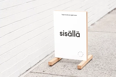 Wooden A-frame sign with white panel displaying black typography reading sisällä on a concrete sidewalk.