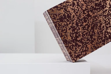 Close-up of a burgundy and copper patterned book balanced on a white pedestal against a neutral studio background