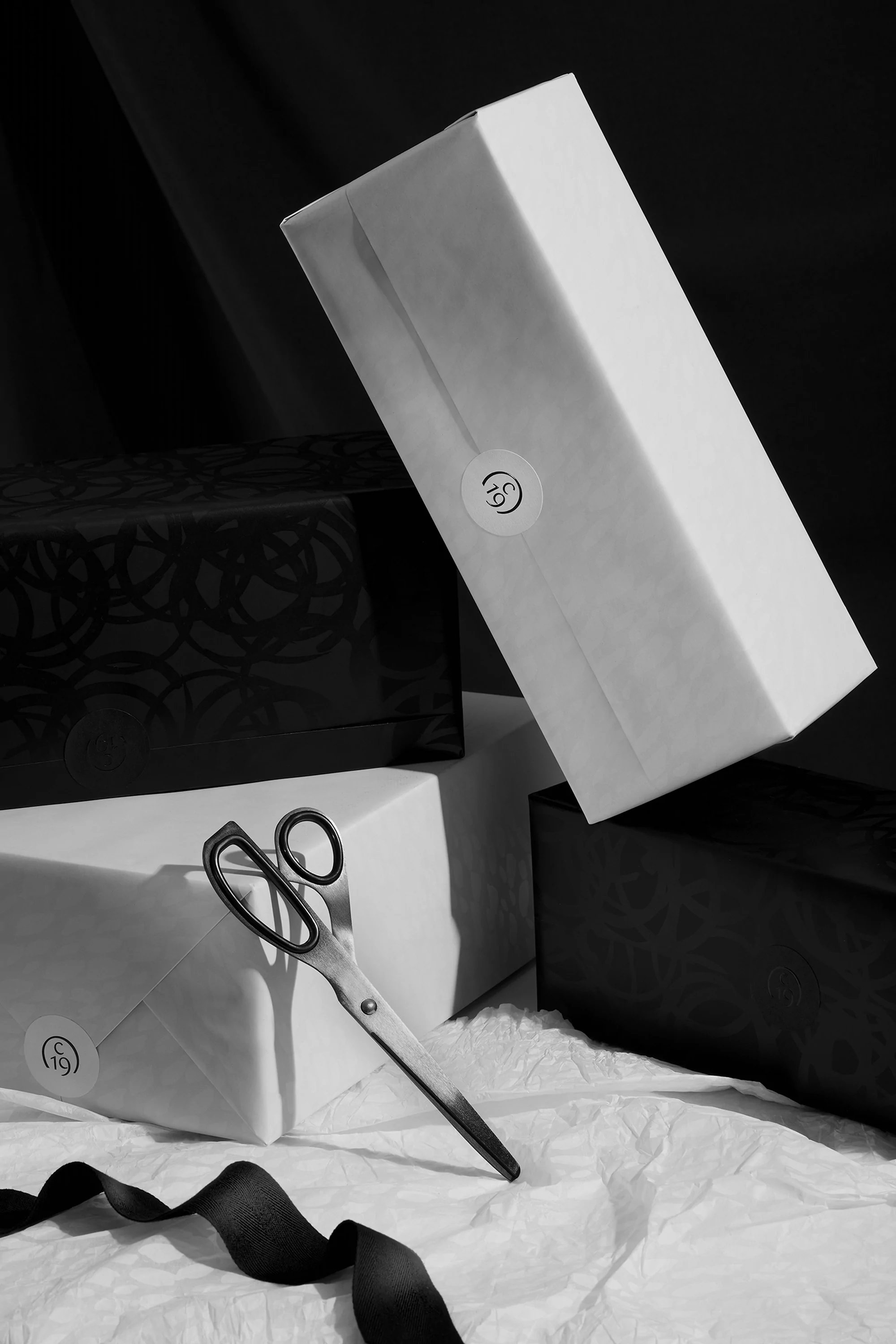Black and white still life featuring stacked gift boxes, a pair of scissors, and ribbon on wrinkled paper in studio lighting.