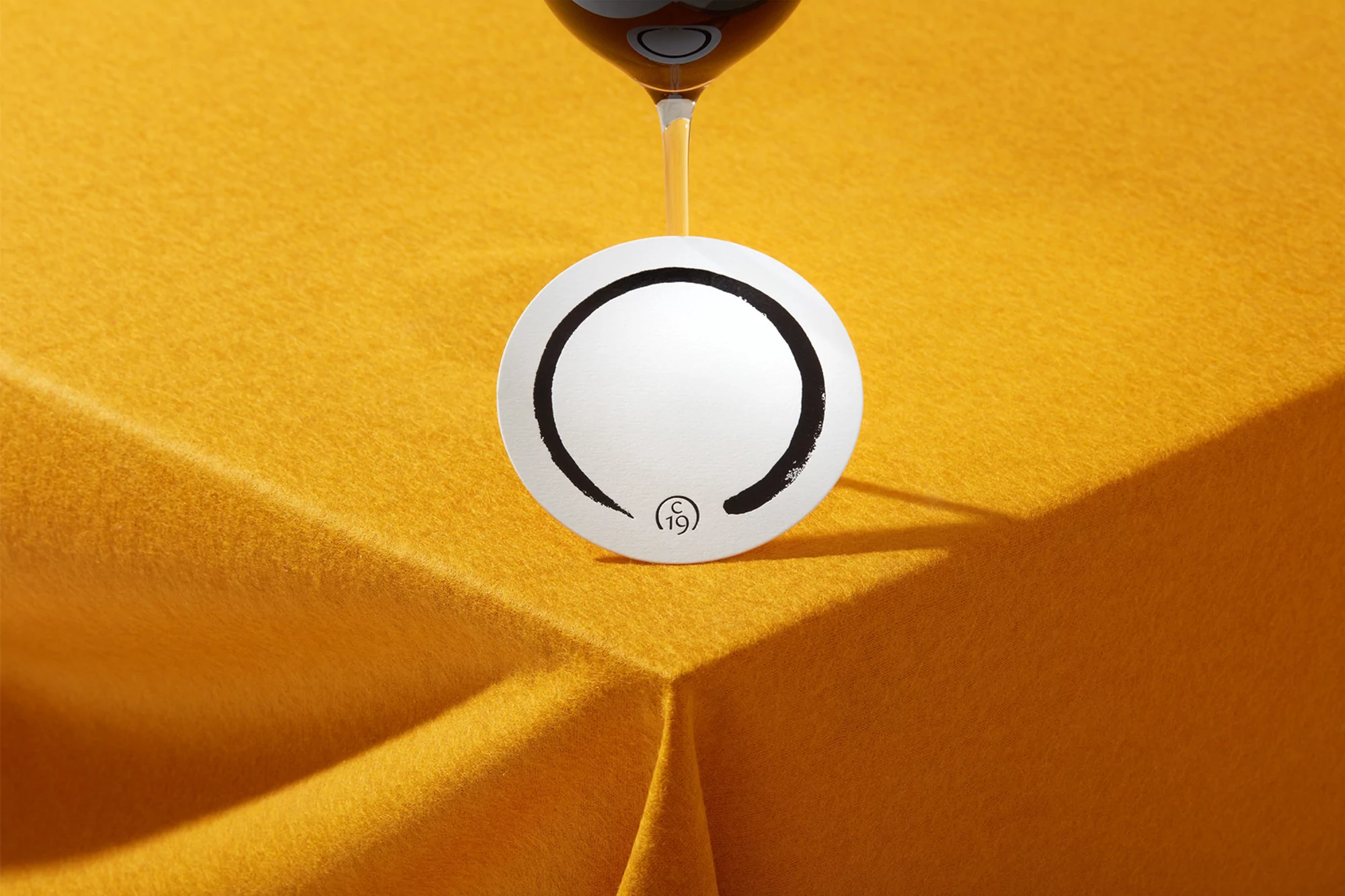 Overhead shot of a wine glass above a coaster with a black and white design on a yellow felt surface, creating a simple and elegant composition.