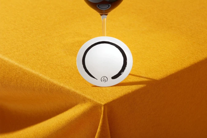 Overhead shot of a wine glass above a coaster with a black and white design on a yellow felt surface, creating a simple and elegant composition.