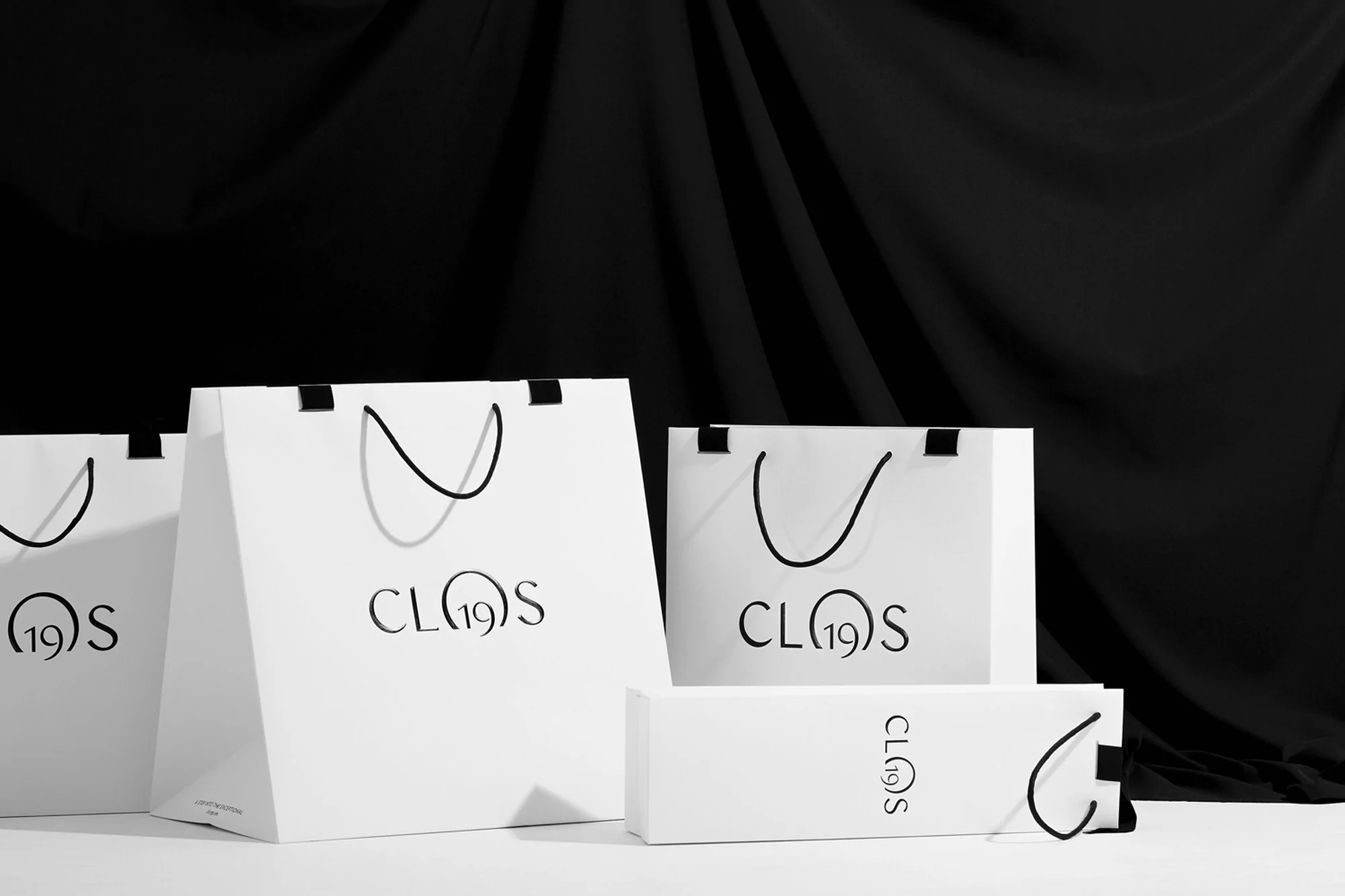 Several white shopping bags and a small rectangular box are grouped together against a dark, draped background in a studio setting.