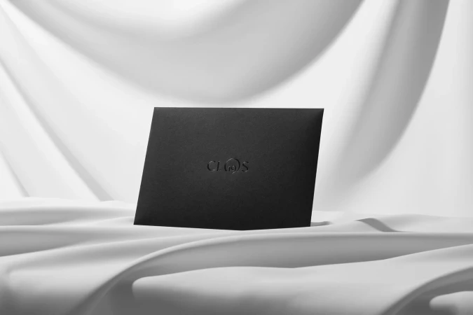 Monochromatic product shot of a black invitation or greeting card with embossed logo on a textured white fabric background in studio lighting.