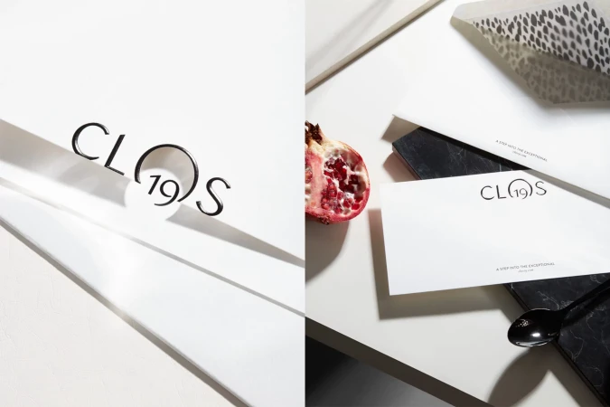 Promotional still life featuring the Clos19 logo, a sliced pomegranate, a black marble coaster, a black spoon, and dappled light.