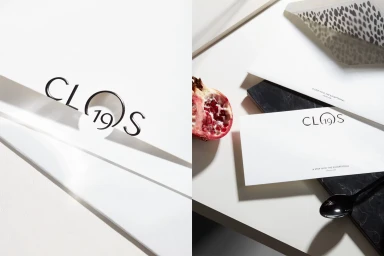 Close-up of branded stationery with the text Clos19, a cut pomegranate half, and a black spoon on white surface