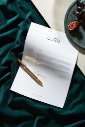 Letterhead stationery sheet with Clos19 logo and gold pen resting on emerald green draped fabric with a dark plate of figs and grapes.