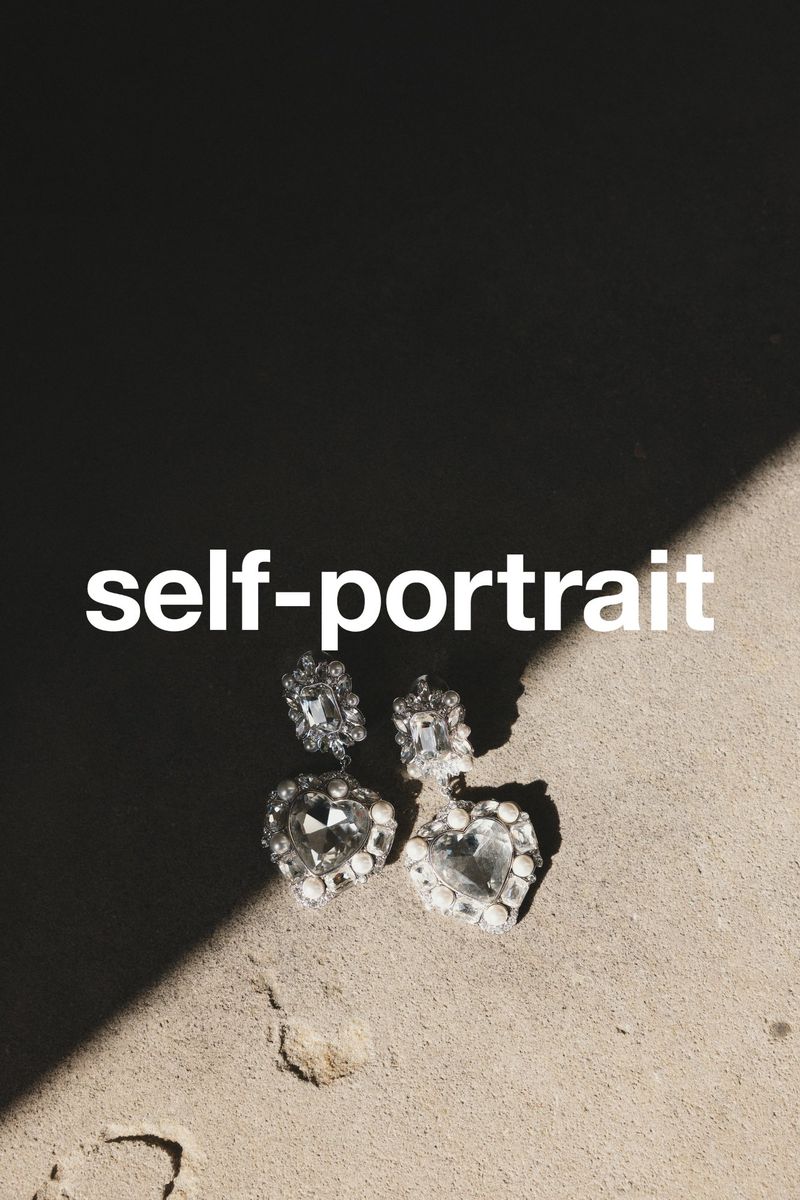 Pair of heart-shaped crystal earrings by Self-Portrait on a textured surface, shadowed on top and illuminated from the bottom.