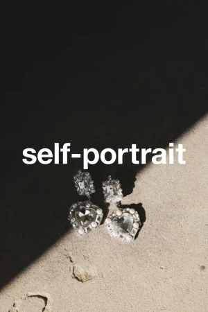 Pair of heart-shaped crystal earrings by Self-Portrait on a textured surface, shadowed on top and illuminated from the bottom.