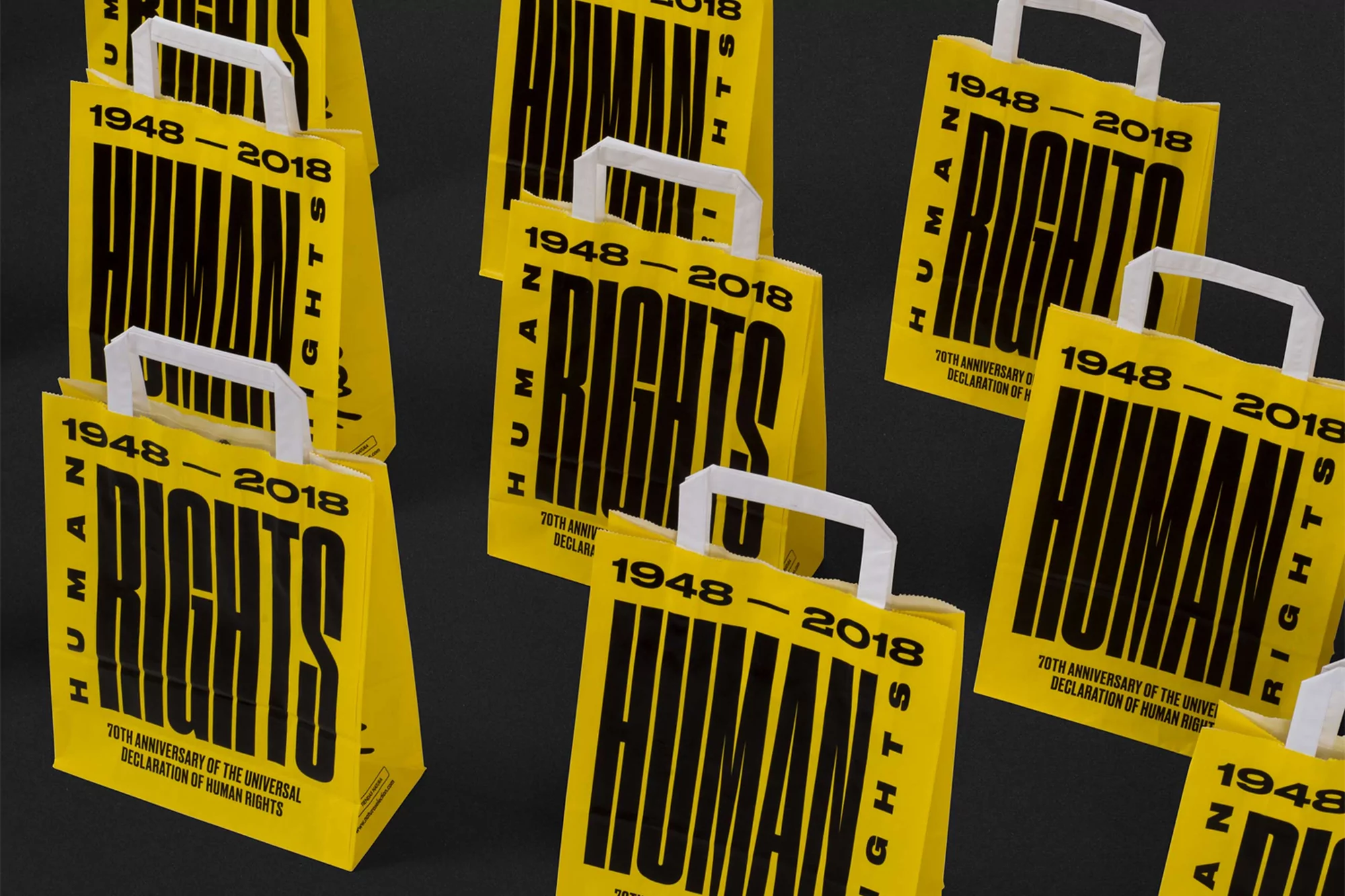 Several yellow paper shopping bags feature 'Human Rights 1948-2018' text and white handles, set against a dark backdrop in a studio shot.