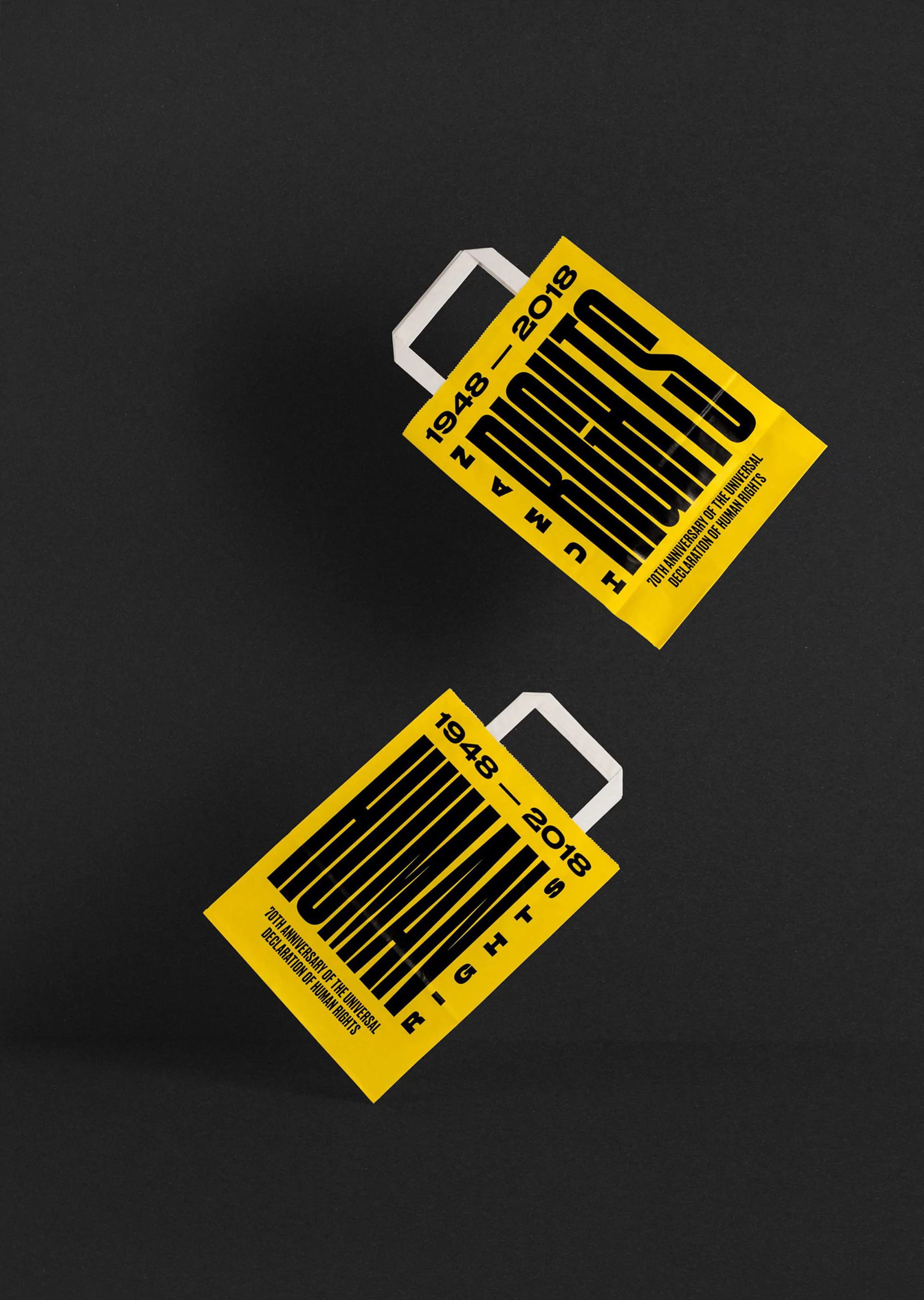 Two yellow shopping bags with 'Human Rights 1948-2018' printed on them, set against a solid dark gray background. Studio lighting.