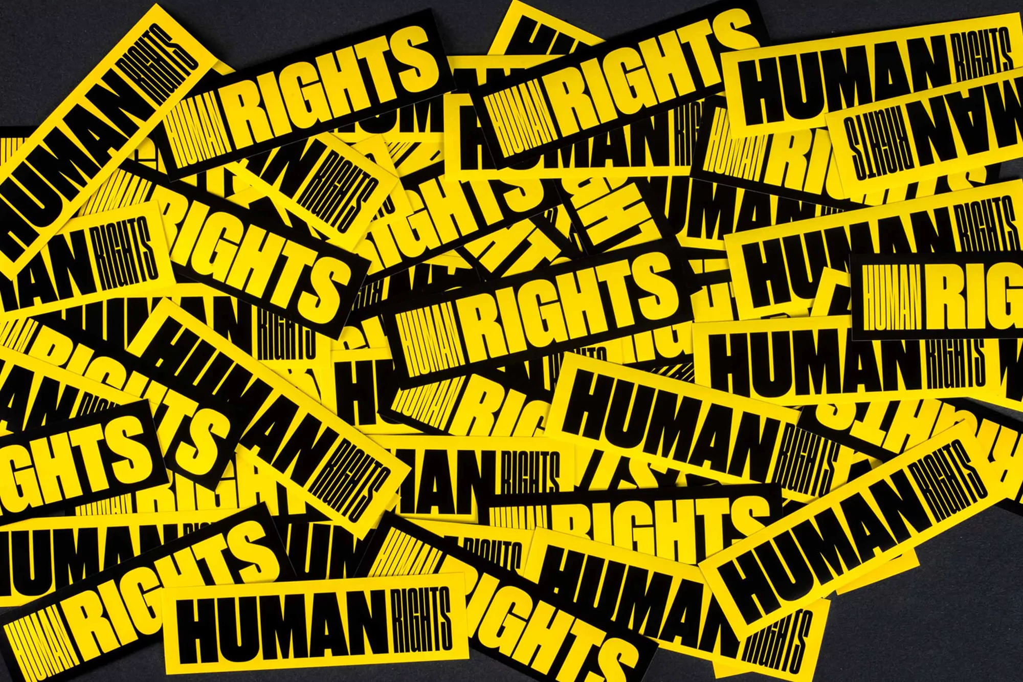 Close-up of overlapping yellow stickers with black text 'Human Rights' in a pile, creating a graphic pattern on a dark gray surface.