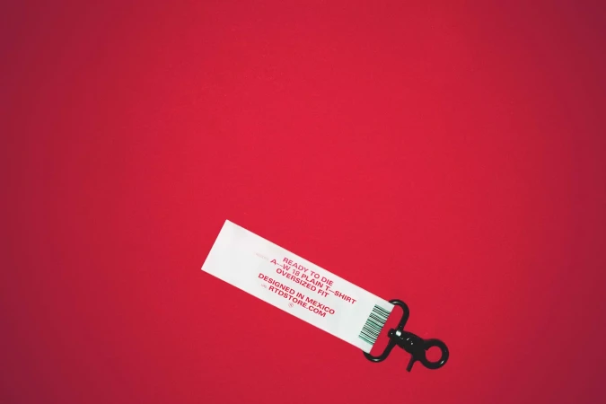 White clothing tag with black lanyard on red background, featuring fashion brand name, product type, and 'Designed in Mexico' text.