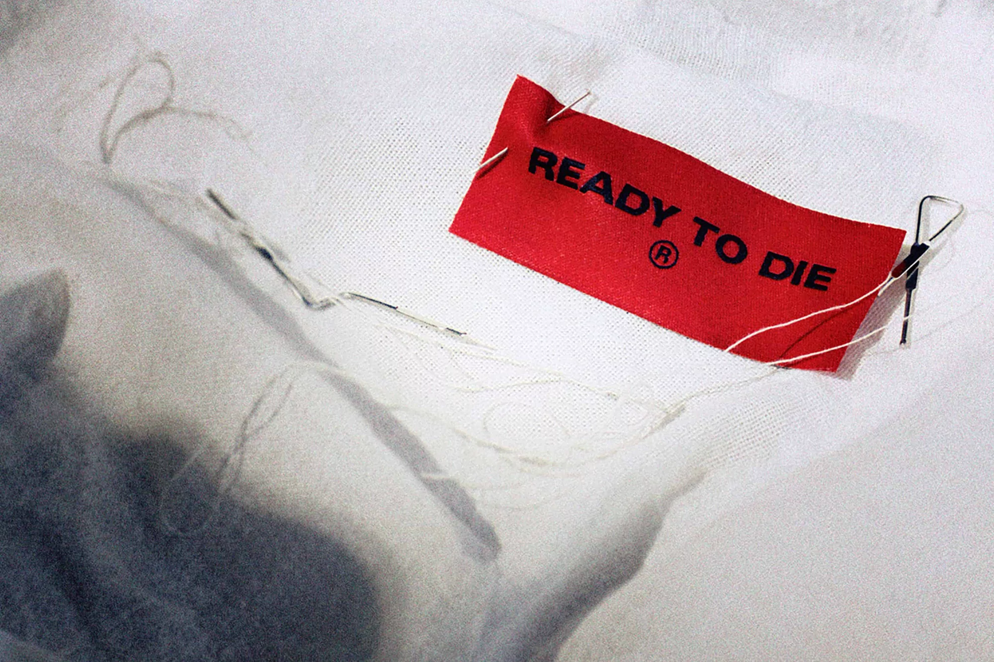 Close-up shot of a red 'Ready to Die' tag attached to a white fabric surface, secured with pins and a metal clip.