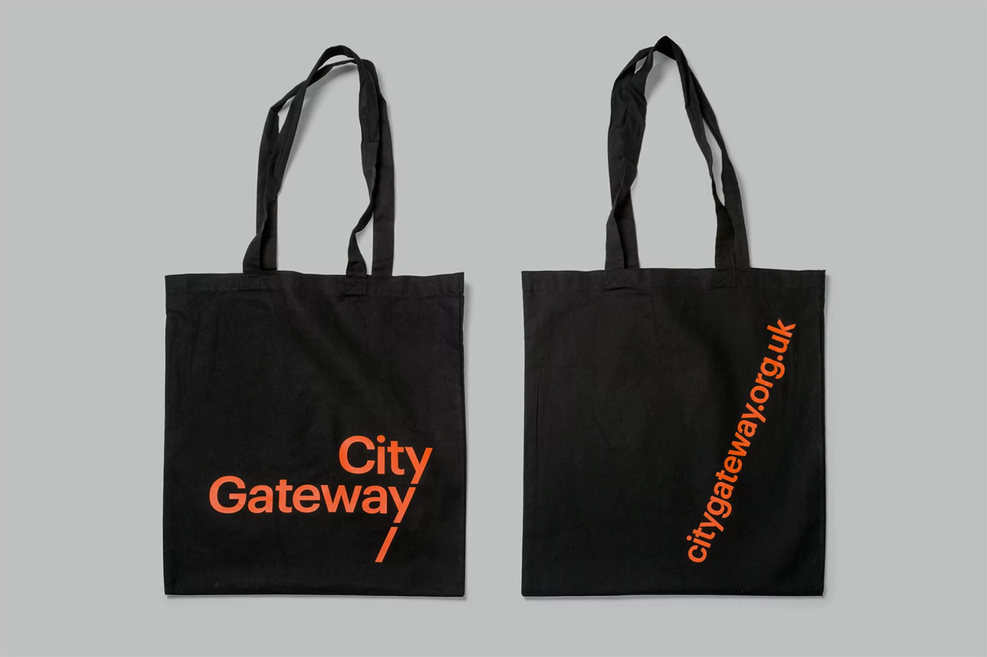 Two black tote bags with orange text on a gray background, one displaying 'City Gateway' and the other 'citygateway.org.uk'.