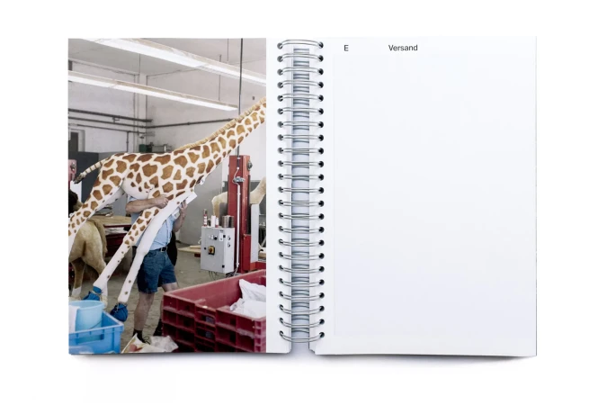 Open notebook displaying a giraffe prop being assembled in an industrial setting with visible workers and equipment, bright overhead lighting.