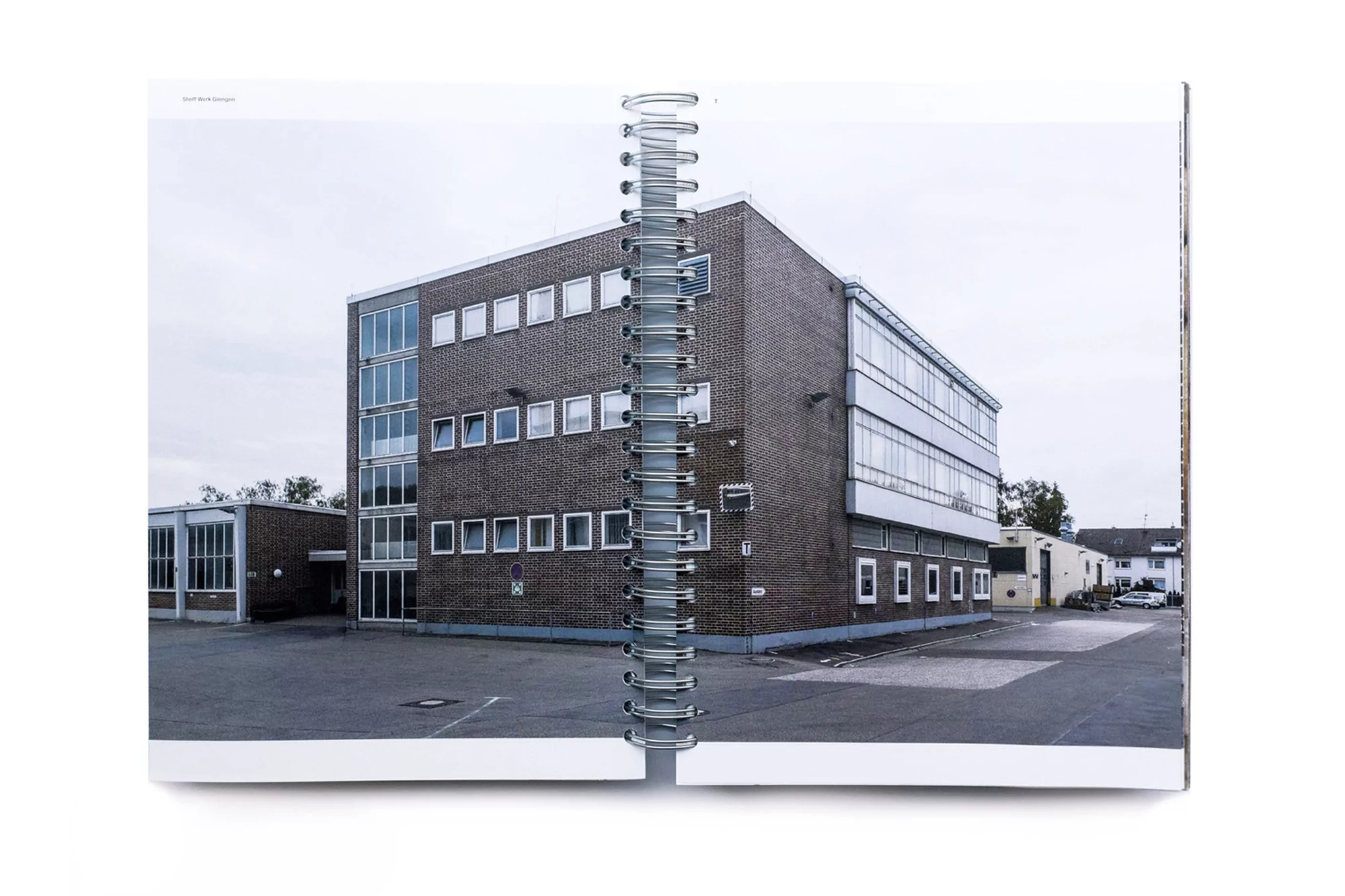 Architectural photograph in a book featuring a brick building with many windows under an overcast sky, next to a paved parking lot.