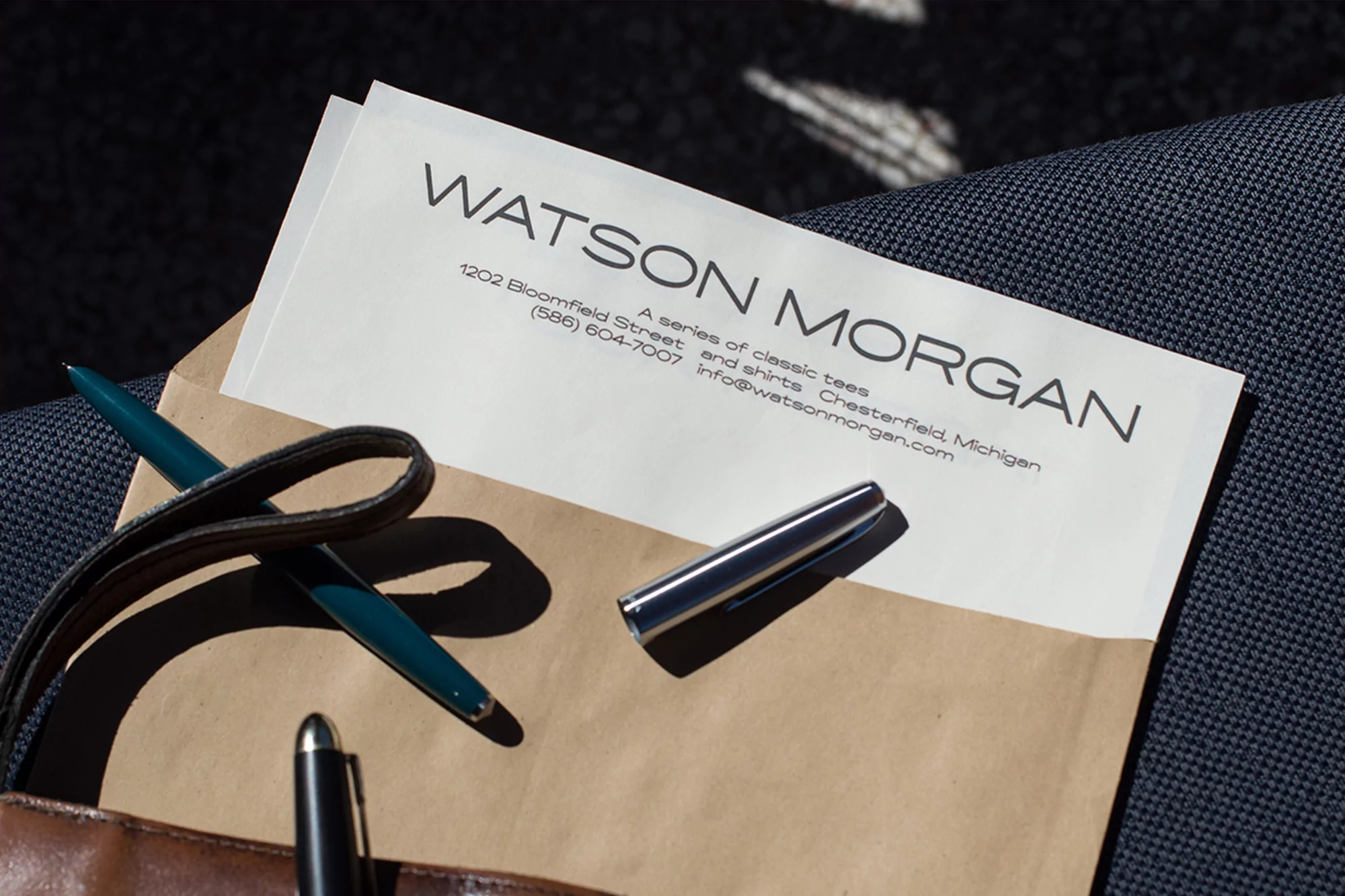 Close-up of a paper with the company name Watson Morgan on top of a brown envelope with a pen set and a navy blue textile.