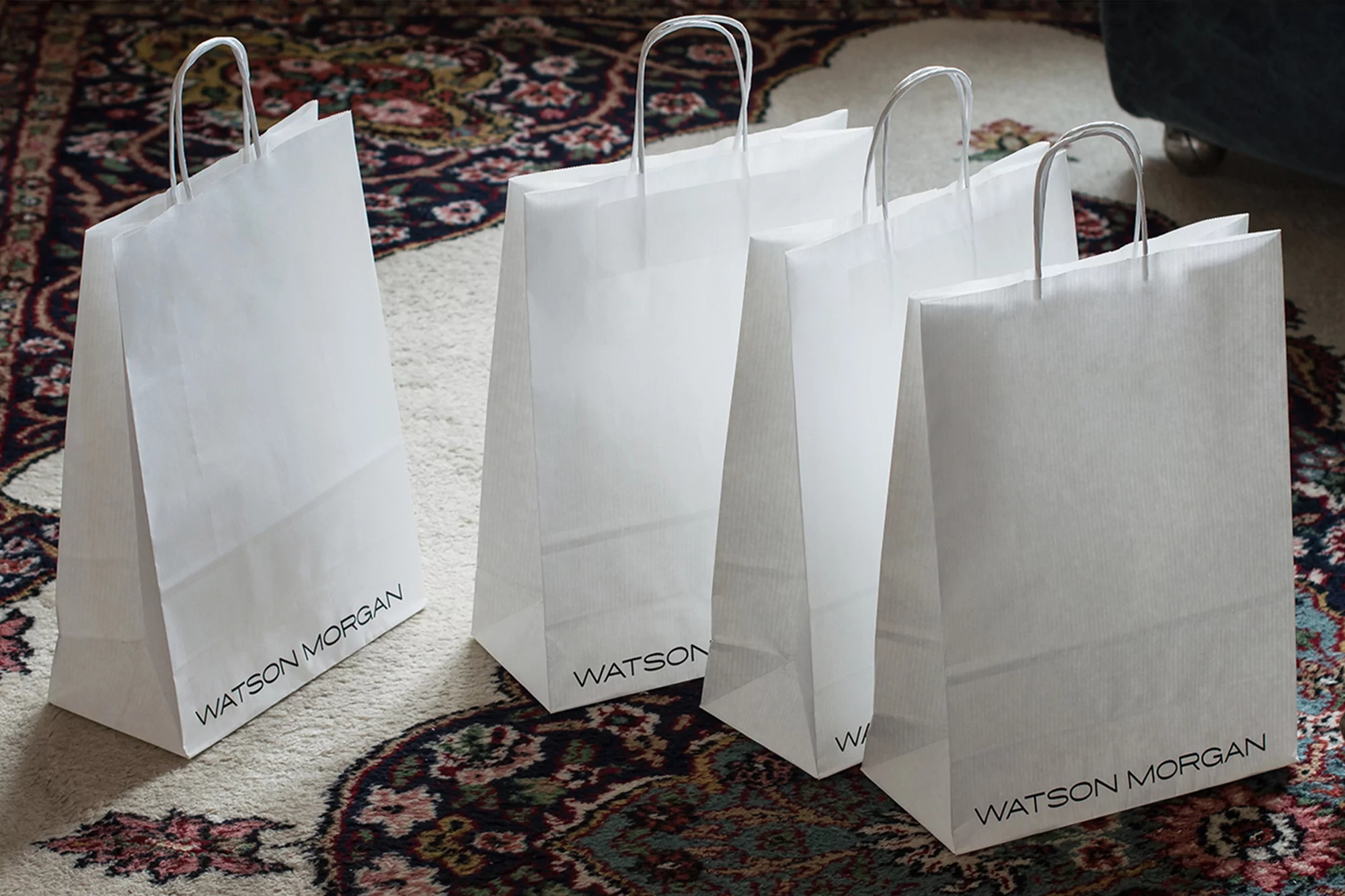 Four white paper shopping bags with handles are standing on a patterned rug. The bags have 'WATSON MORGAN' printed on them.