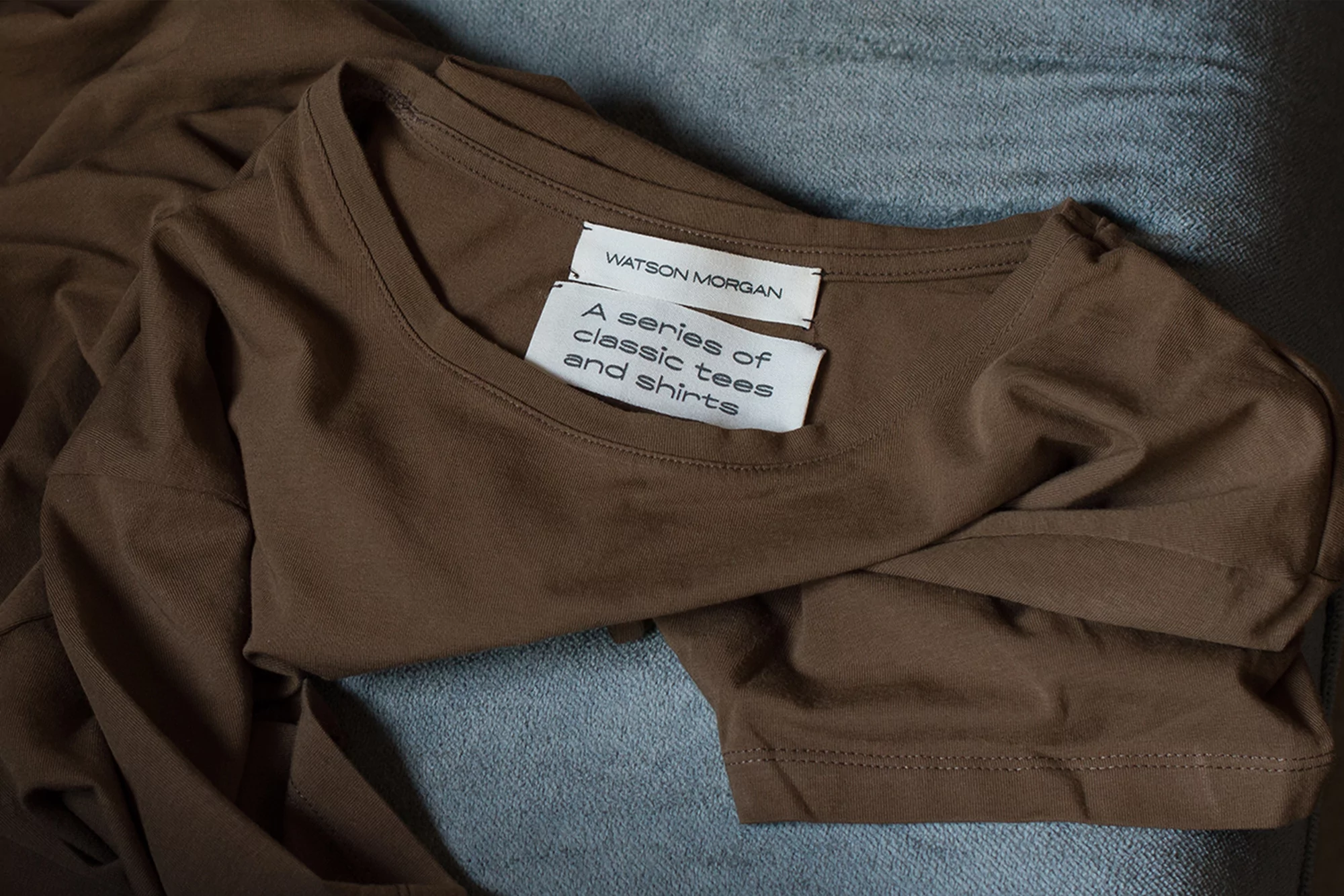 Close-up of a folded brown t-shirt with a label, showcasing a simple design and soft fabric texture against a blue textile backdrop.