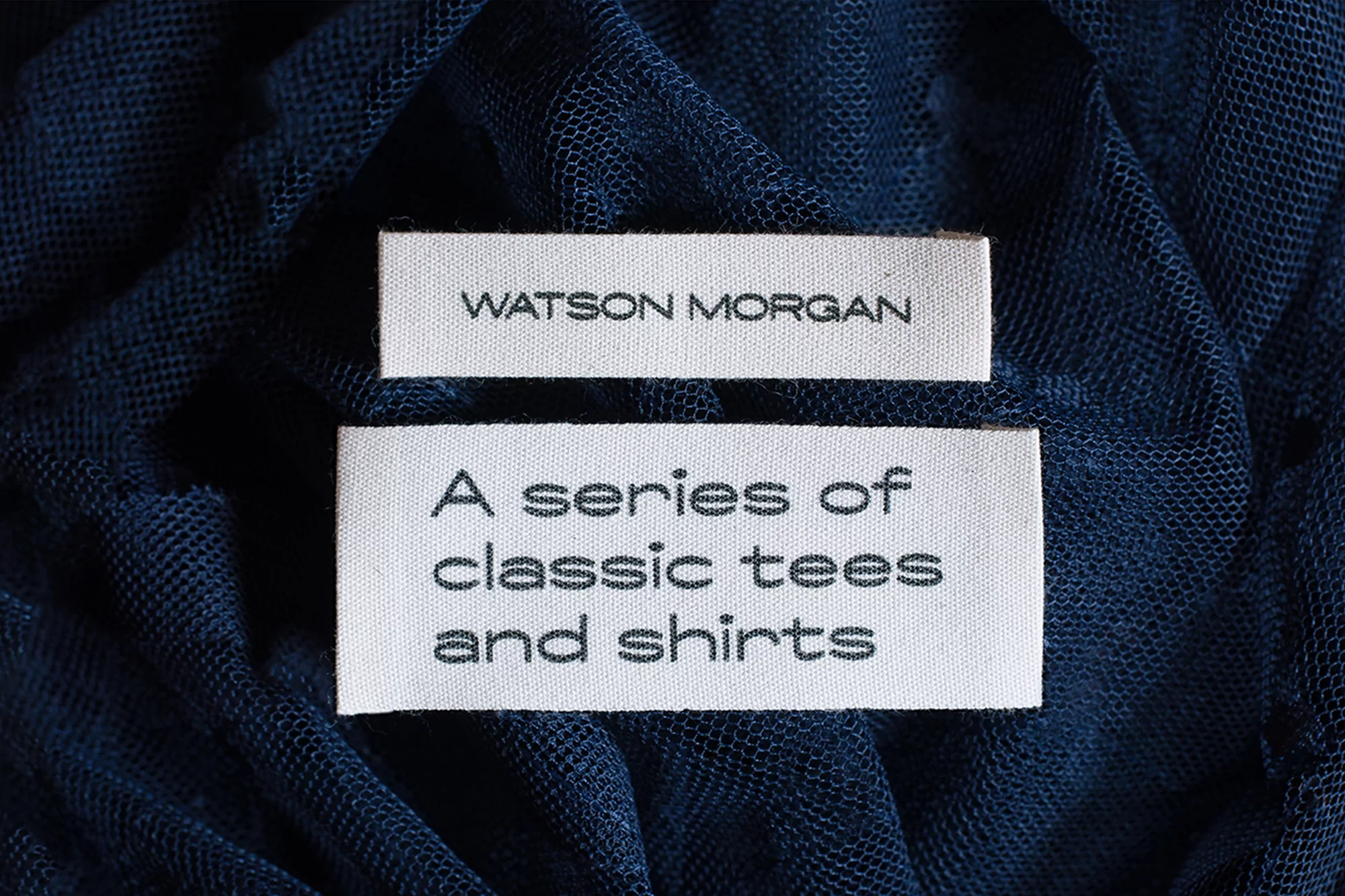 Two white fabric labels with black text sit atop a textured navy blue textile, showcasing a clothing brand or product description.