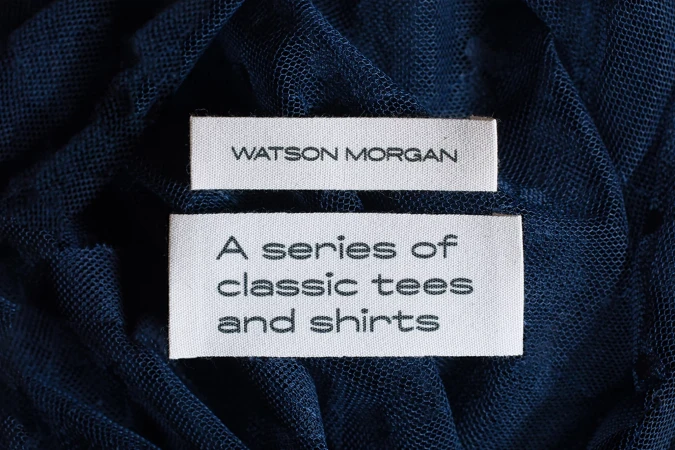 Two white fabric labels with black text sit atop a textured navy blue textile, showcasing a clothing brand or product description.