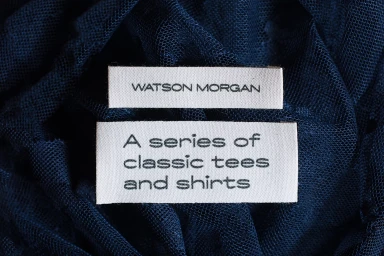 Two woven clothing labels reading Watson Morgan and A series of classic tees and shirts resting on deep blue textured fabric.