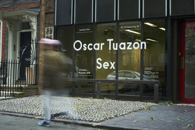A person walks by the Oscar Tuazon Sex exhibition at Jonathan Viner gallery in London, with large window display and gray cobblestone walkway.